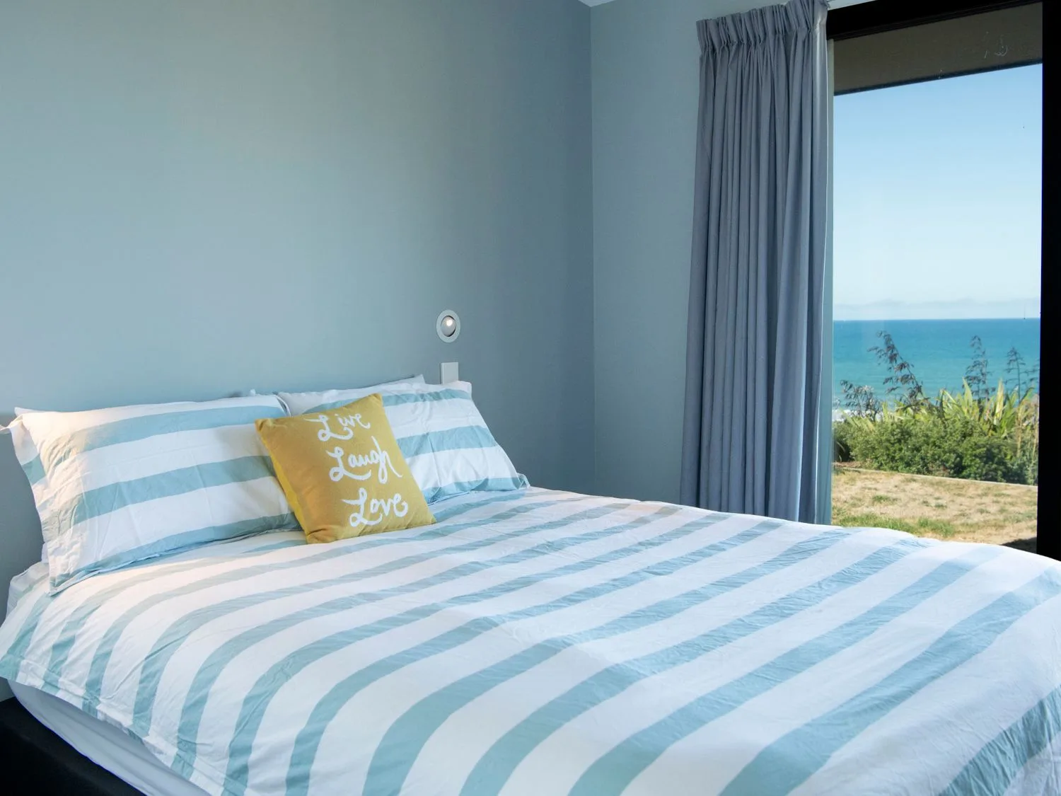 A bedroom with striped bedding and a yellow pillow beside a window overlooking the sea.