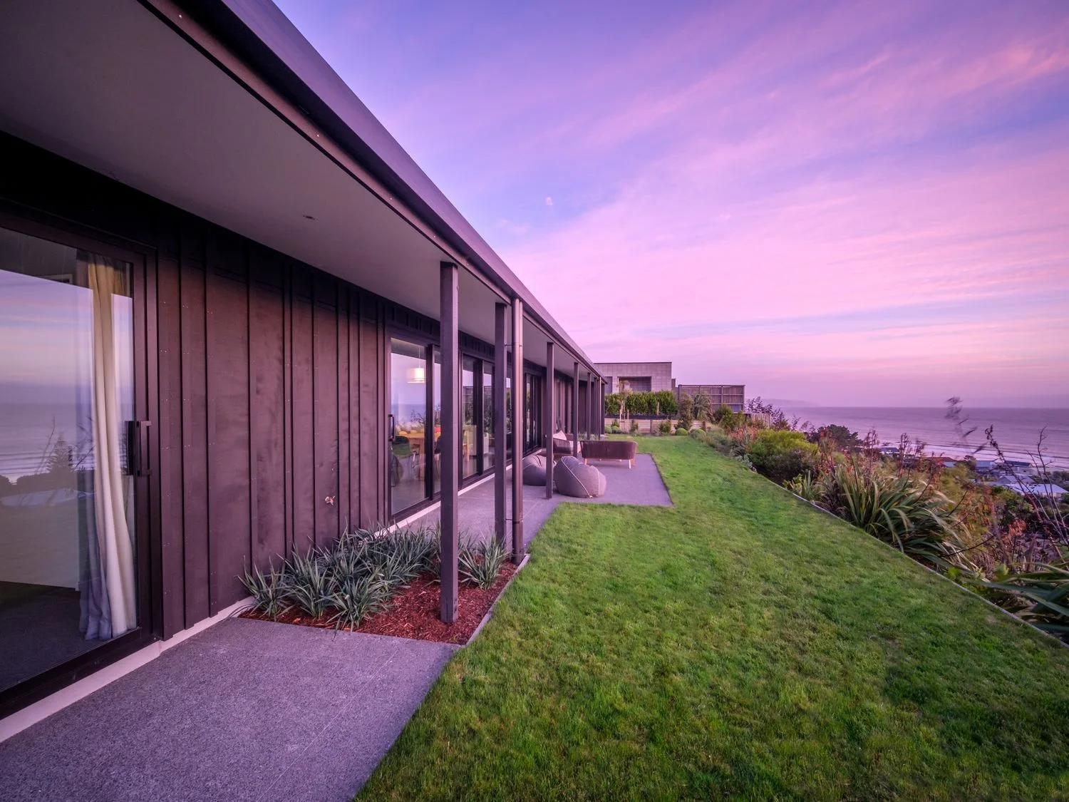 A modern house opens onto a grassy lawn beside the ocean at sunset.