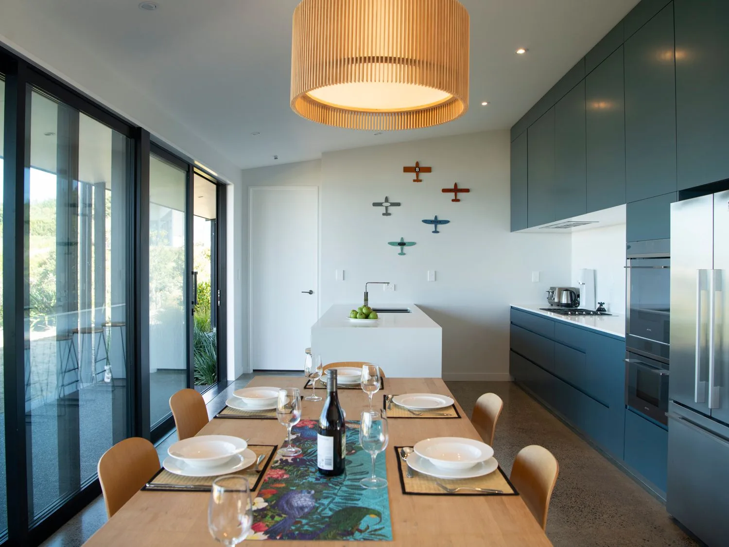 A kitchen with a wooden dining table set for six next to large sliding glass doors and blue cabinetry at Lightning Ridge Views - Waimarama Holiday Home in Waimarama