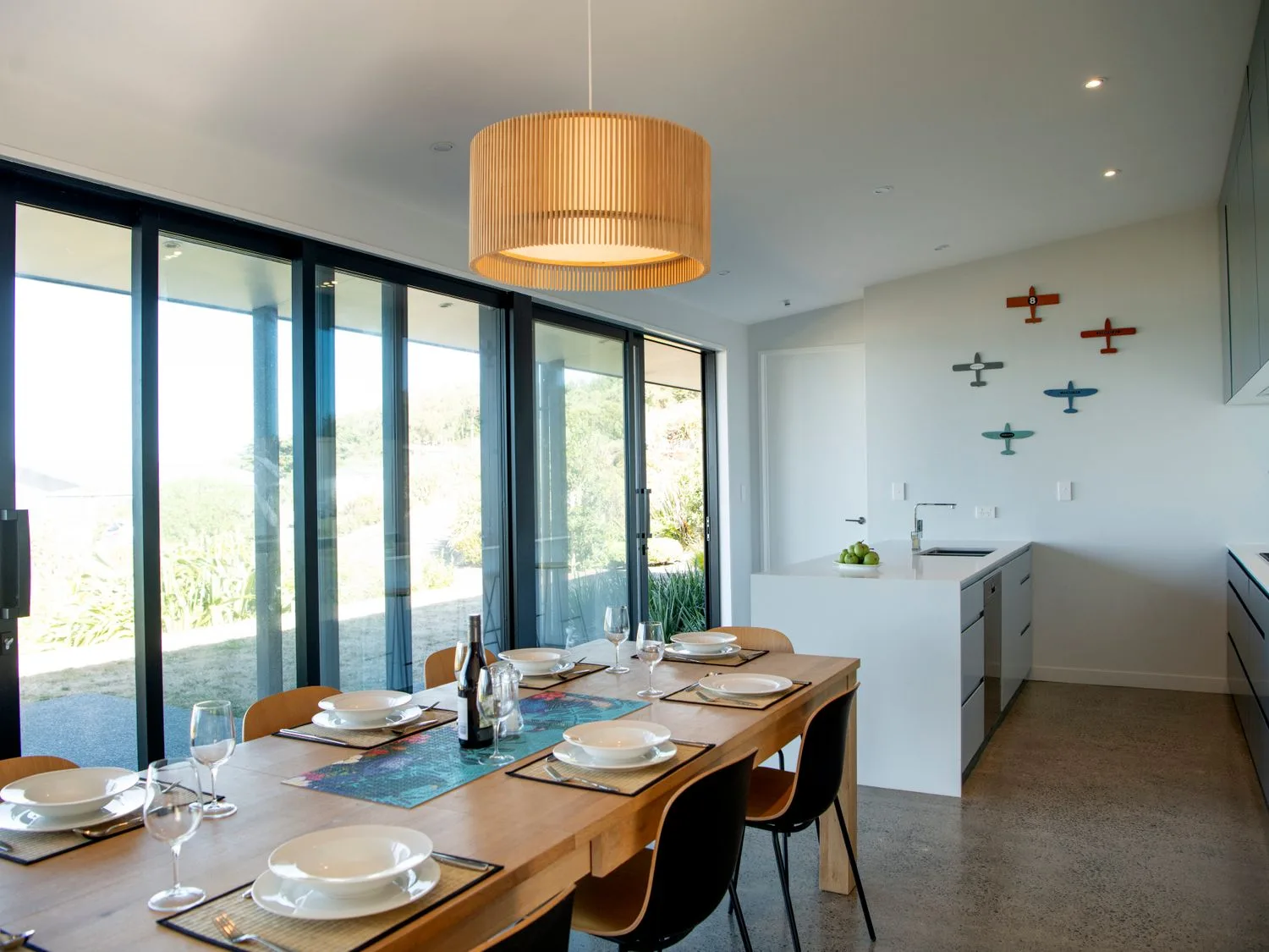 A dining area with a wooden table set with plates and glasses next to a kitchen with a white island and wall decor at Lightning Ridge Views - Waimarama Holiday Home in Waimarama
