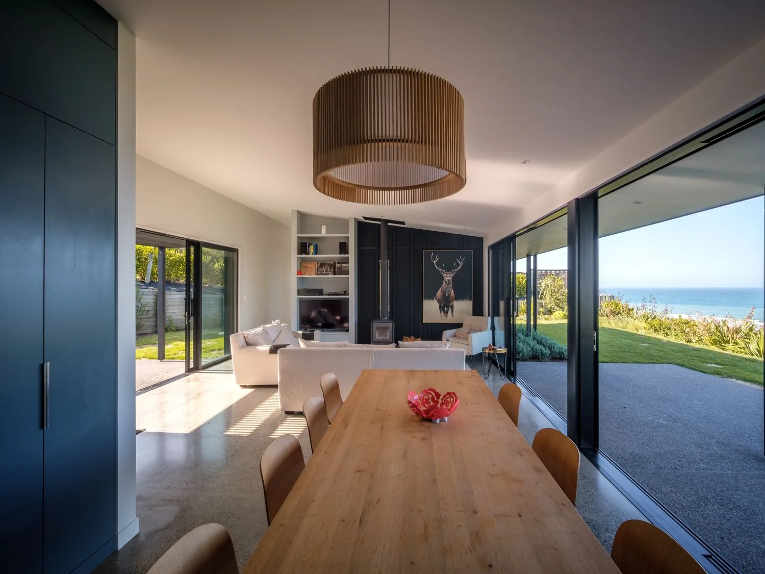 Una zona de comedor con mesa y sillas de madera junto a una sala de estar con sofás y una gran ventana con vistas al océano en Lightning Ridge Views - Waimarama Holiday Home, en Waimarama