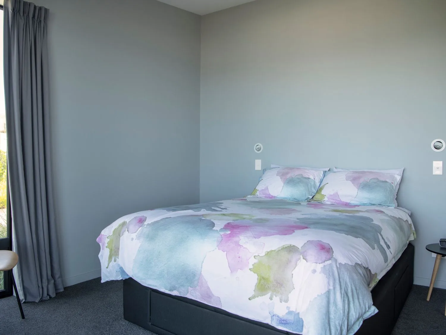 A bedroom with a bed covered in pastel-colored bedding beside a curtained window.
