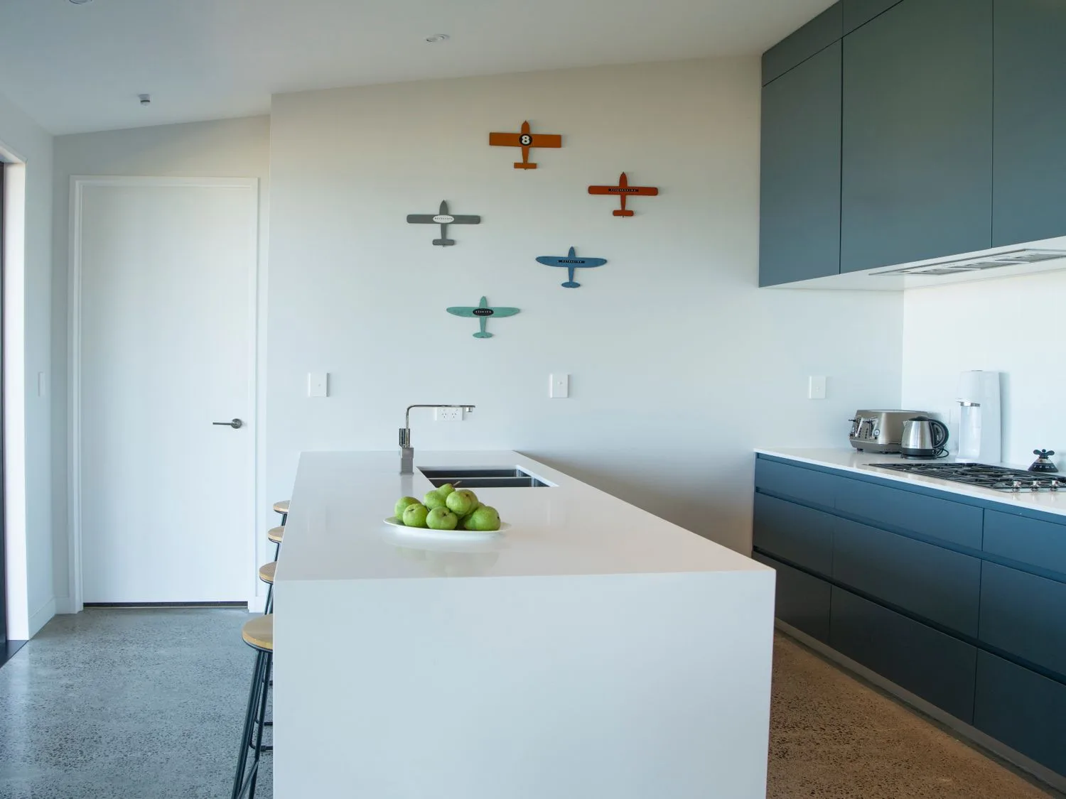 A kitchen with a white island counter with a bowl of green apples stools and blue cabinets on the wall at Lightning Ridge Views - Waimarama Holiday Home in Waimarama