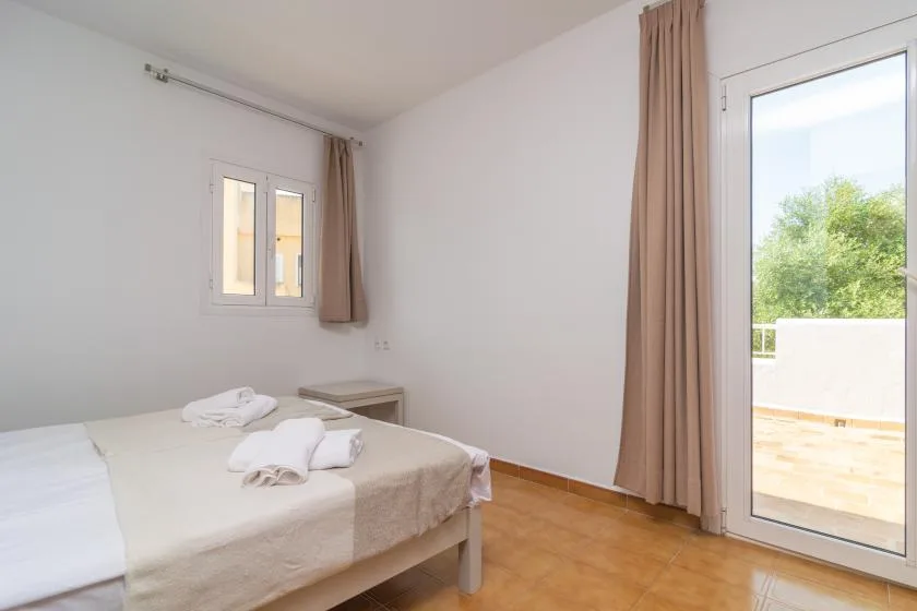 A bright bedroom with a bed beside a curtained glass door opening onto a balcony.