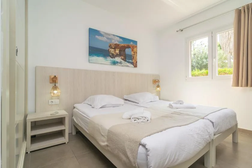 A bright bedroom with two beds and a window overlooking greenery.