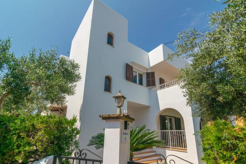 A white stucco building with arched windows stands among leafy trees.
