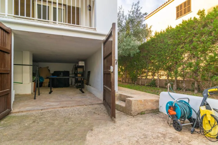 A covered garage area opens onto a paved outdoor space beside a hedge.