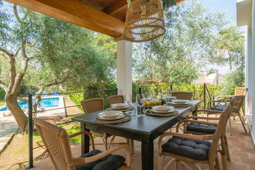 A covered outdoor dining area with a table and cushioned seating overlooks a garden.