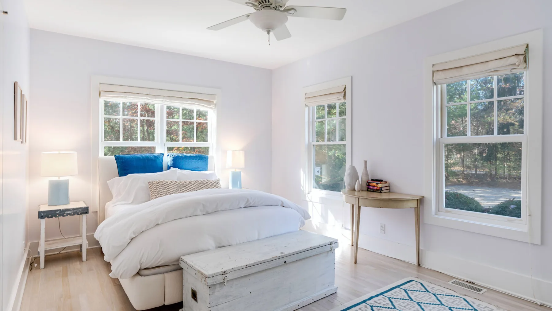 Un dormitorio luminoso con una cama, un baúl blanco y ventanas con vistas a la vegetación.