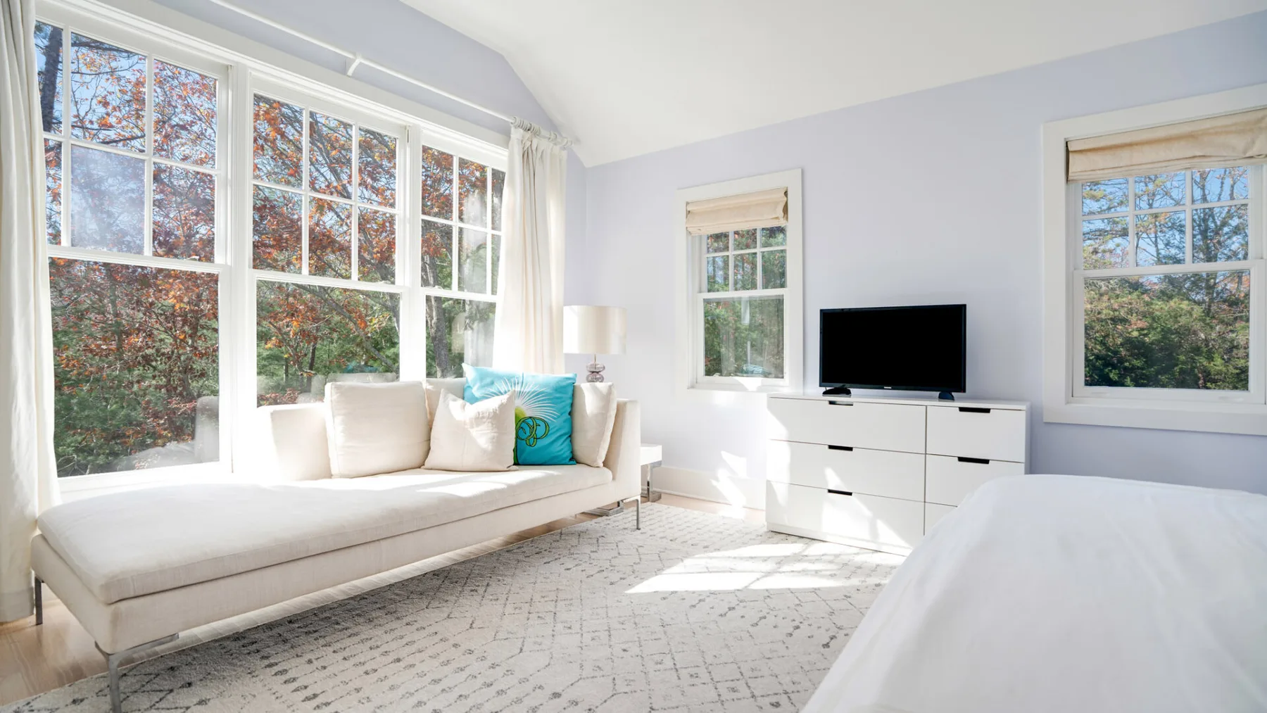 Un dormitorio luminoso con una cama, una zona de estar junto a la ventana y un televisor.