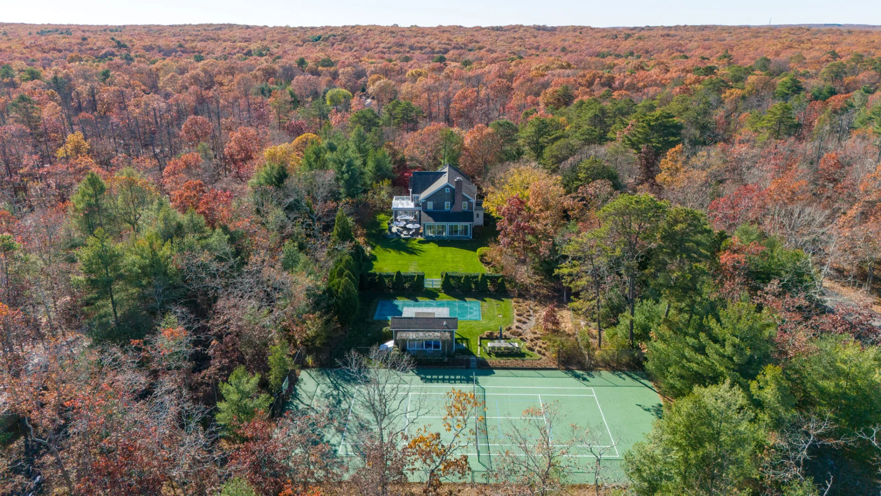 Una vista aérea muestra una casa y una cancha de tenis rodeadas de bosque otoñal.