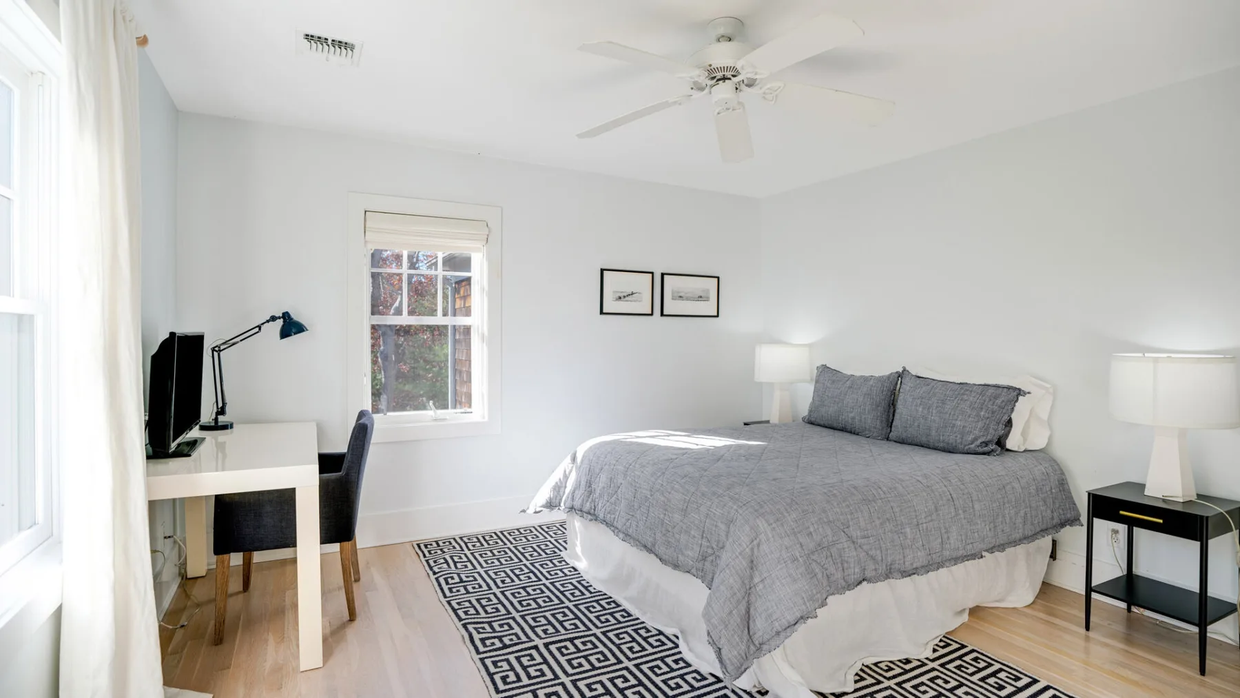 Un dormitorio luminoso con una cama gris y un escritorio junto a la ventana.