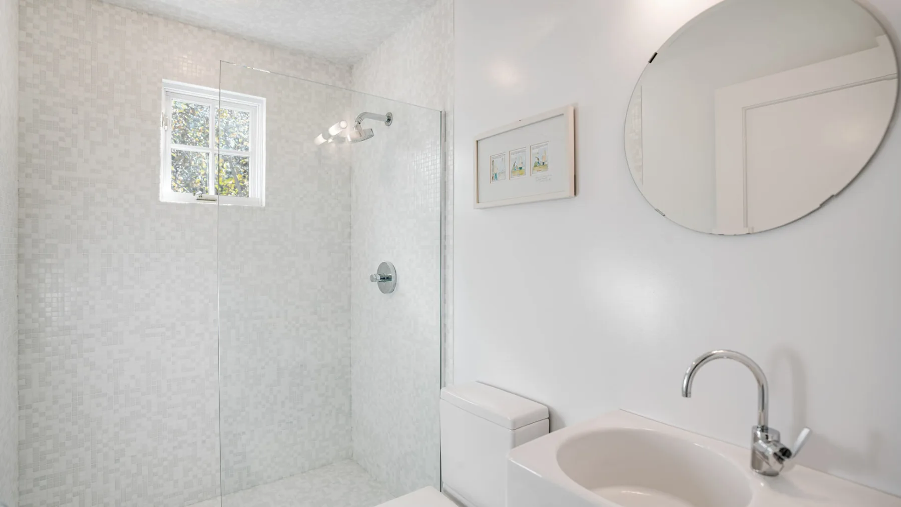 Baño luminoso con ducha de vidrio y lavabo bajo un espejo redondo.