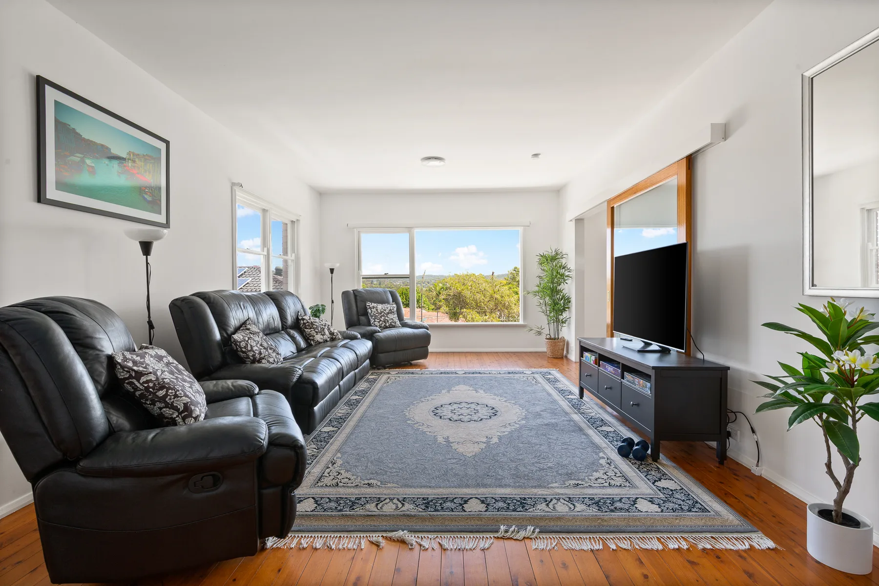 A bright living room has seating, a large rug, and windows overlooking greenery.