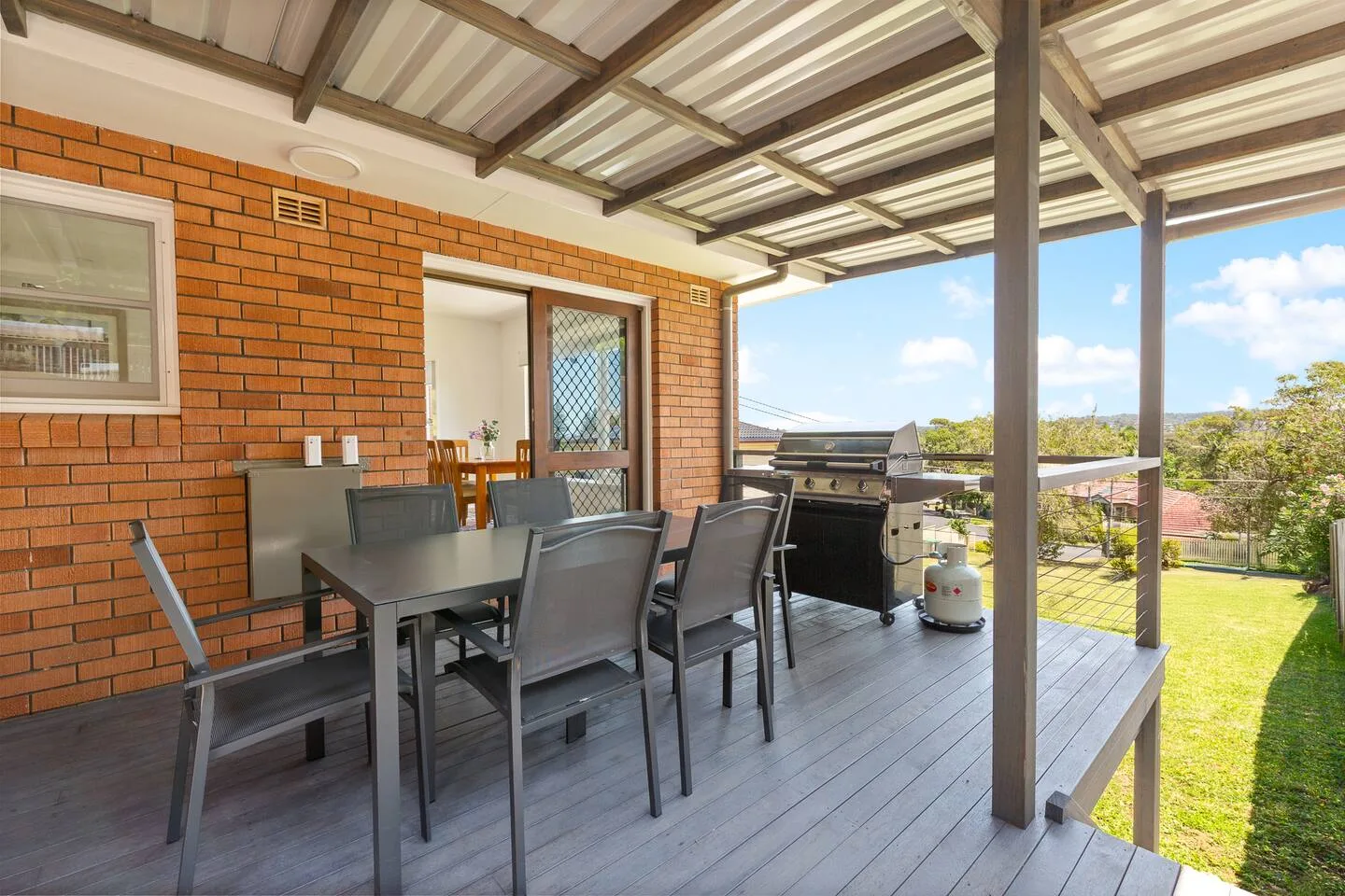 A covered outdoor dining area has a table and seating beside a barbecue.