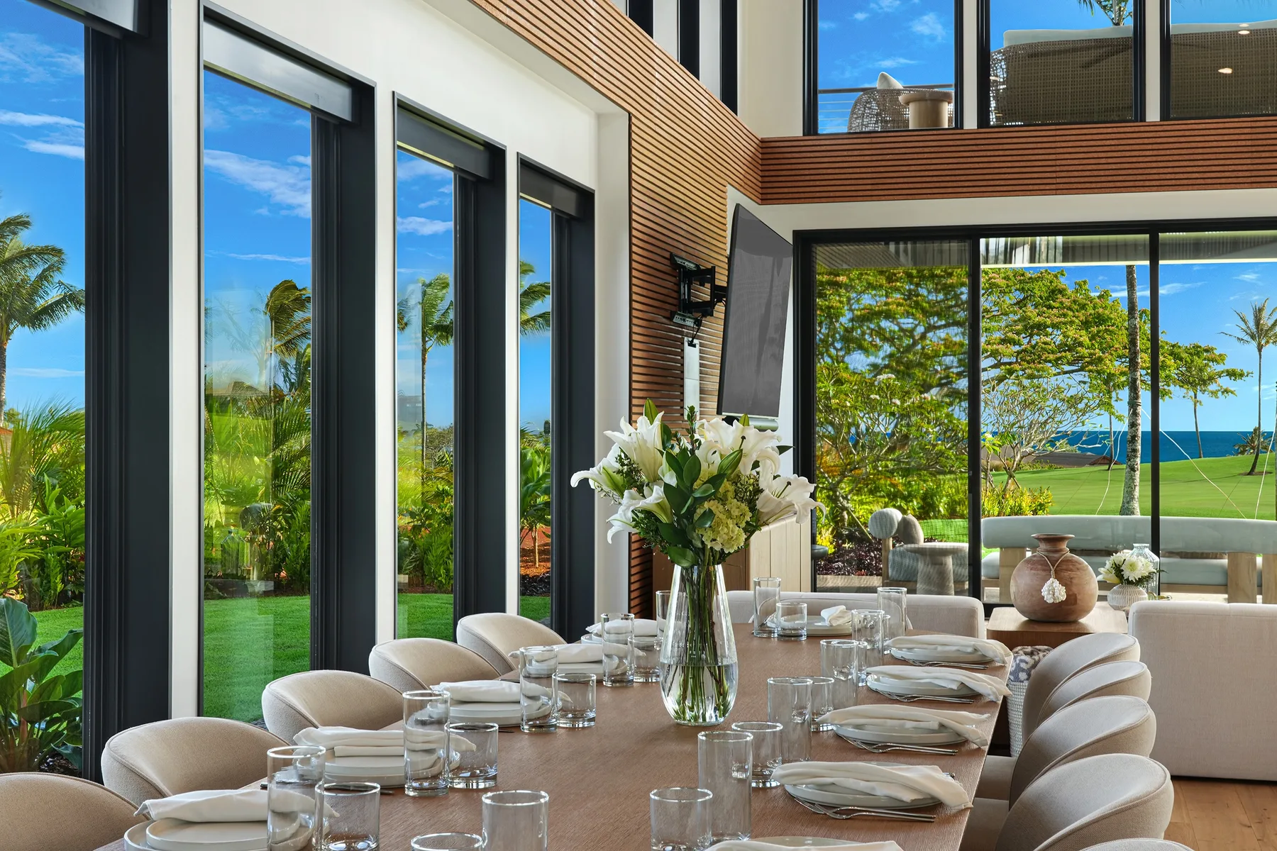 Una mesa de comedor está preparada con cristalería y flores junto a amplios ventanales con vistas a la vegetación y al mar.
