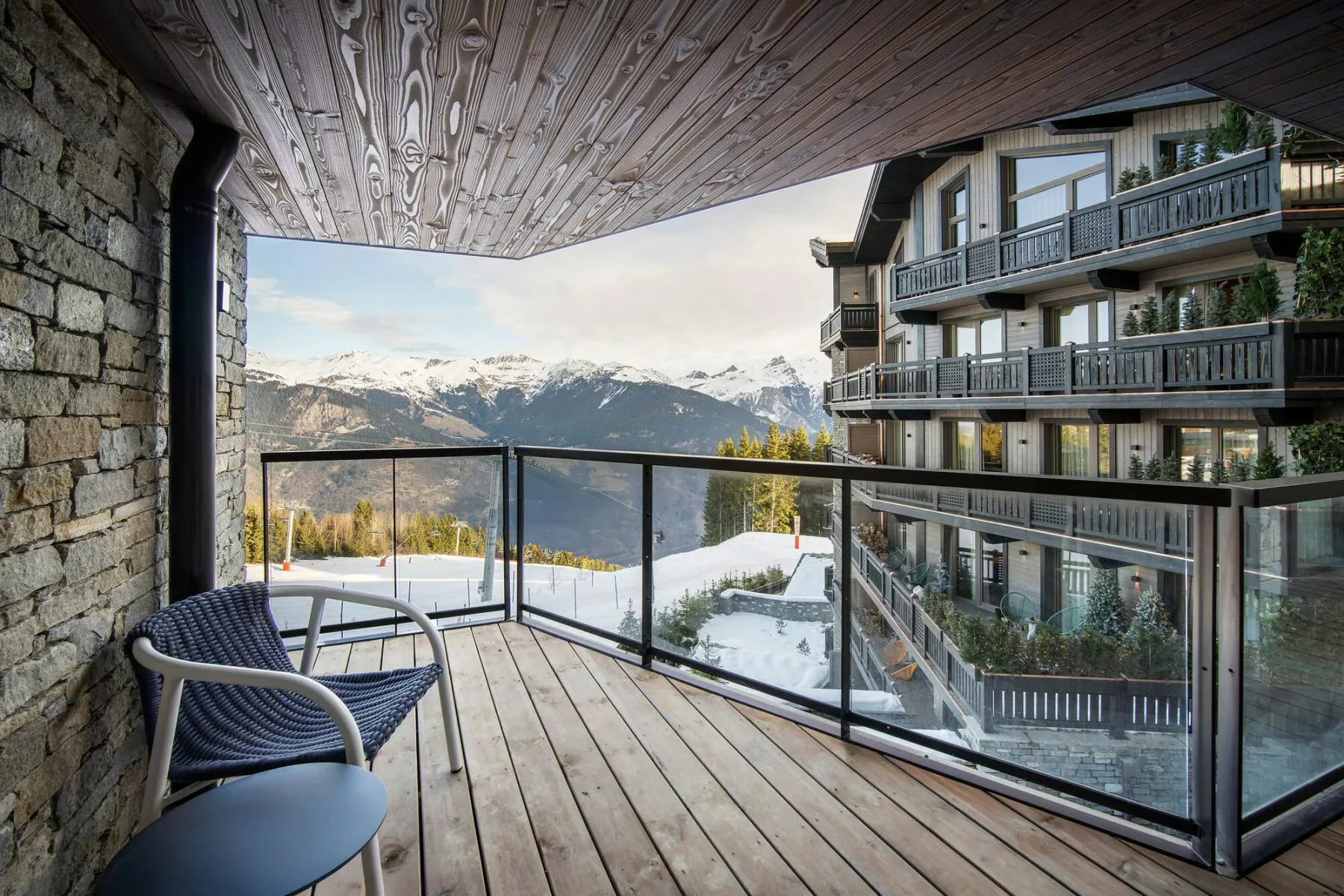 A covered balcony with outdoor seating overlooks snowy mountains and a neighboring building.