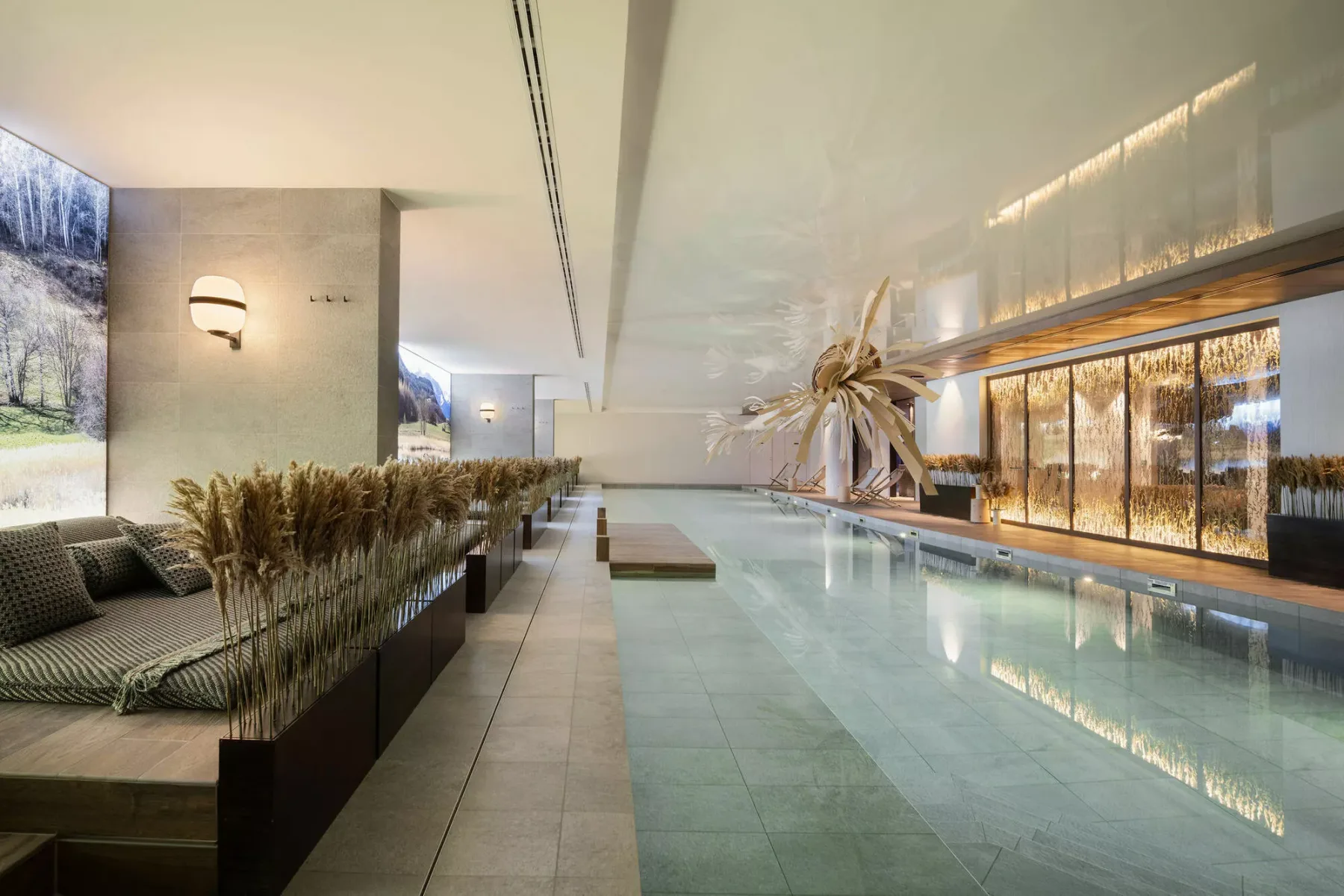An indoor swimming pool runs alongside lounge seating and tall windows.