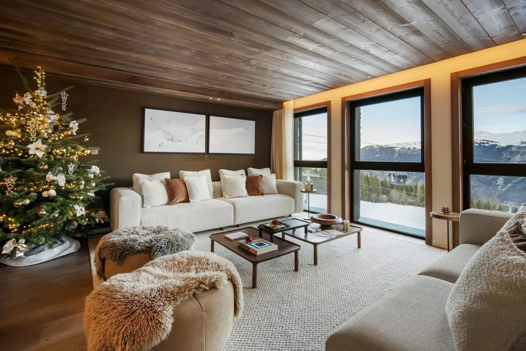 A living room with light seating, a decorated tree, and large windows overlooking snowy mountains.