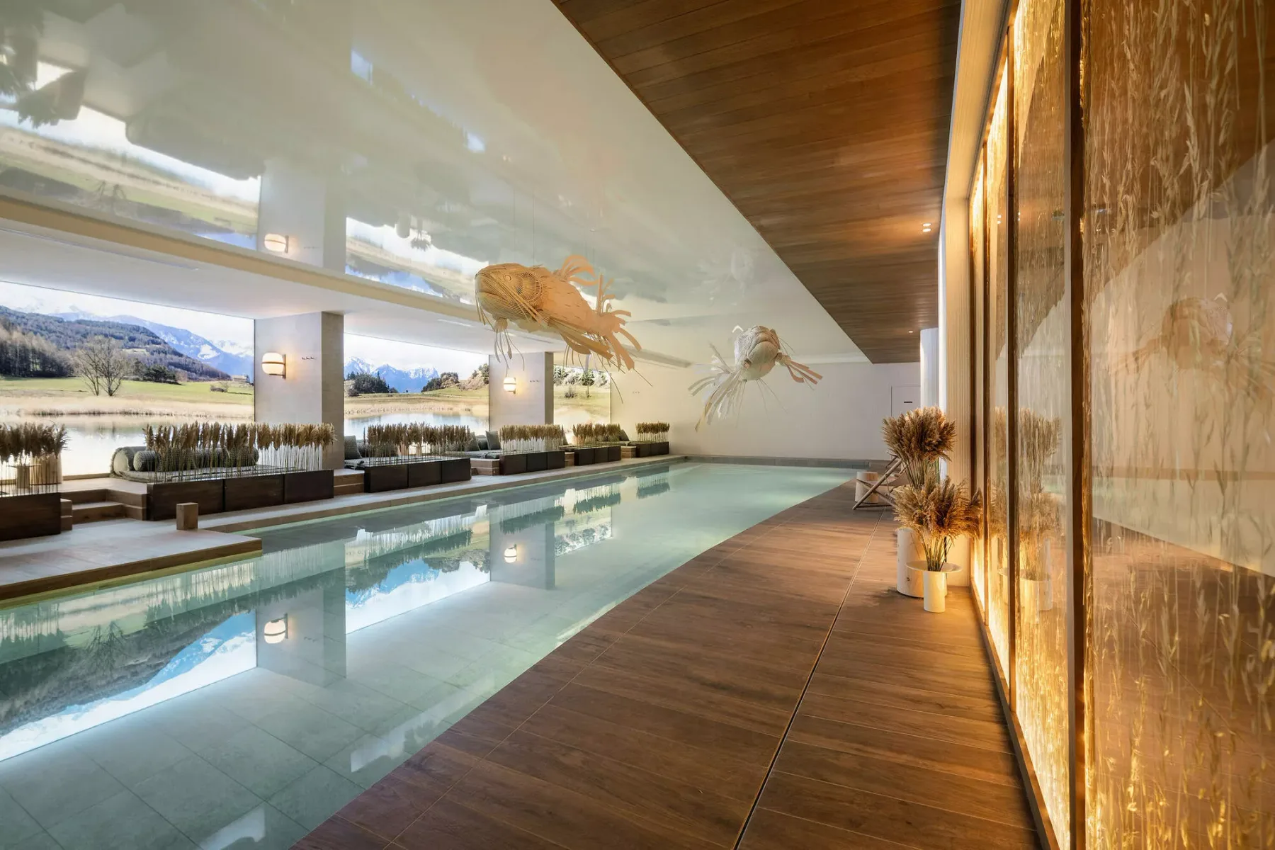 An indoor pool beside a wooden walkway, with large windows overlooking a lake.