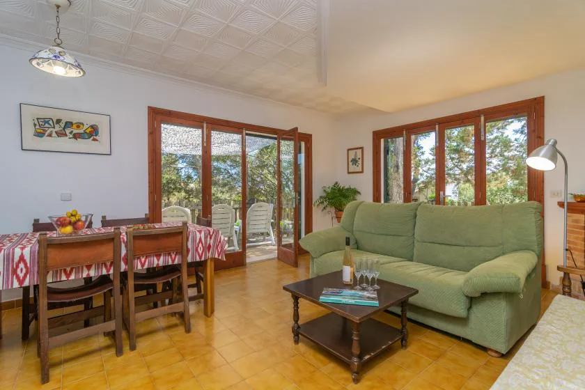 A living and dining room with green seating and large windows overlooking greenery.