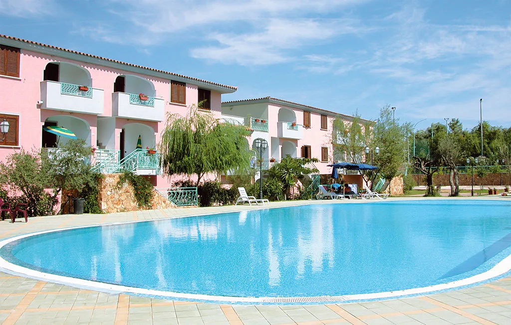 An outdoor swimming pool in front of pink resort buildings and surrounding greenery.