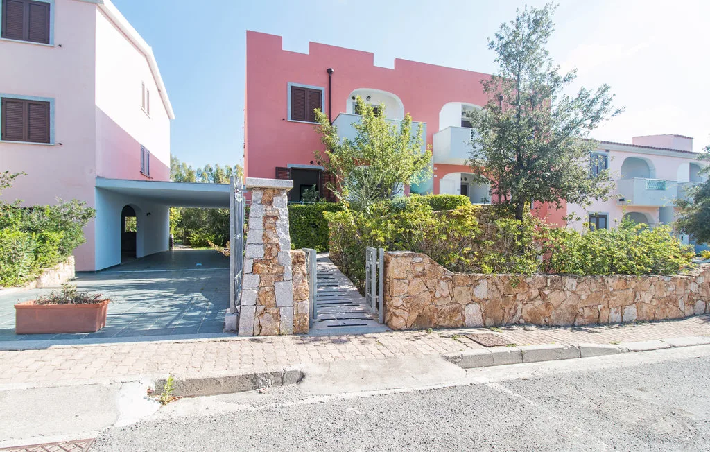 A pink building stands behind a stone wall, with a gated walkway and greenery.