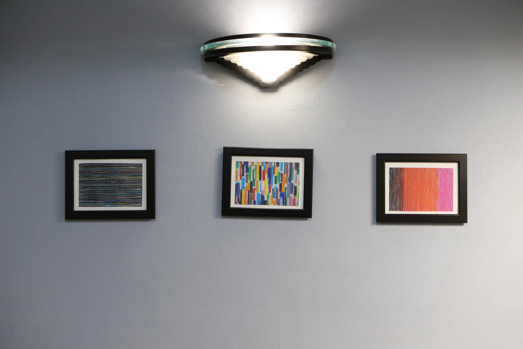 Tres obras abstractas enmarcadas cuelgan de una pared bajo una lámpara.