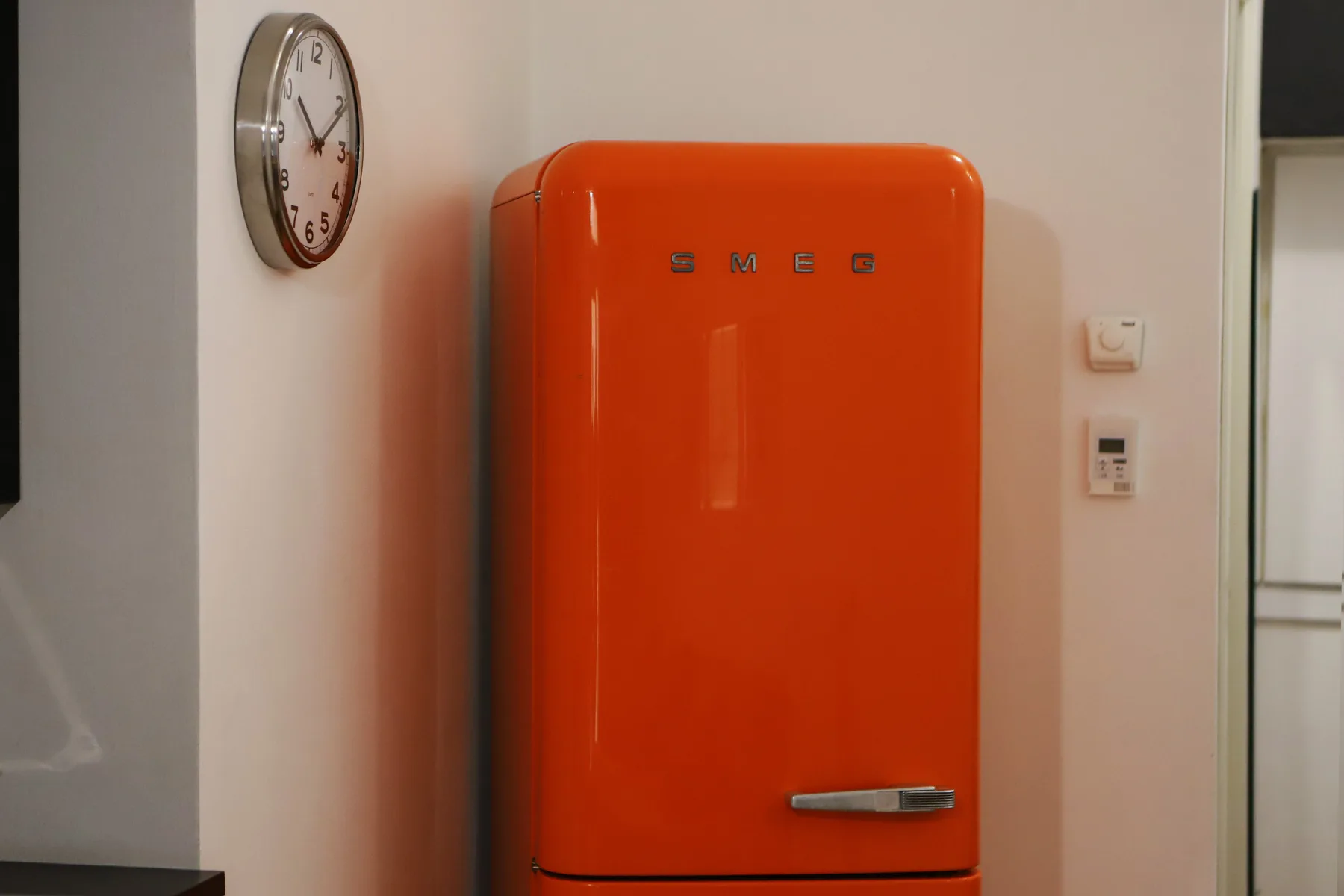 Un refrigerador rojo brillante está junto a una pared clara, debajo de un reloj redondo.