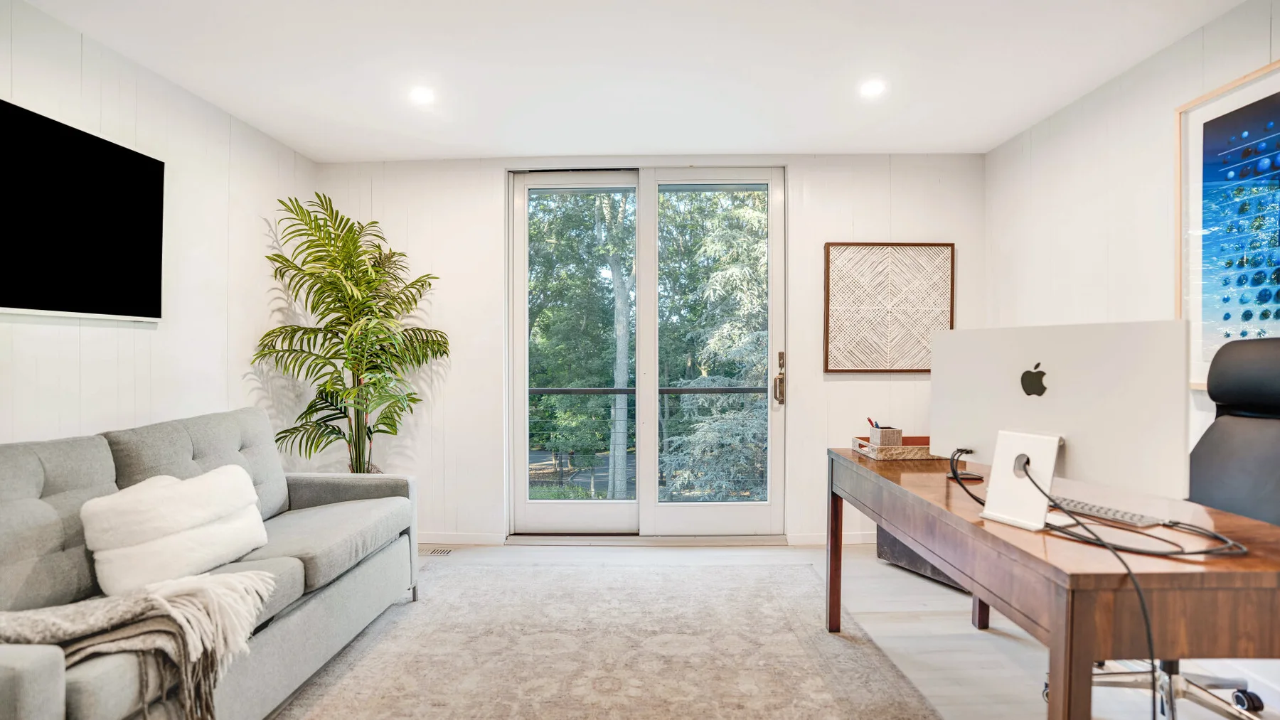 Una sala luminosa con un sofá gris, un escritorio de madera y puertas de vidrio.