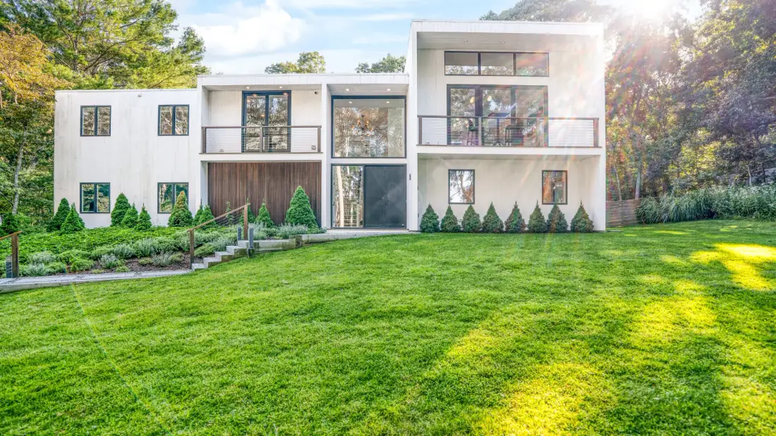 Una casa blanca moderna se alza detrás de un amplio césped verde y un jardín ajardinado.