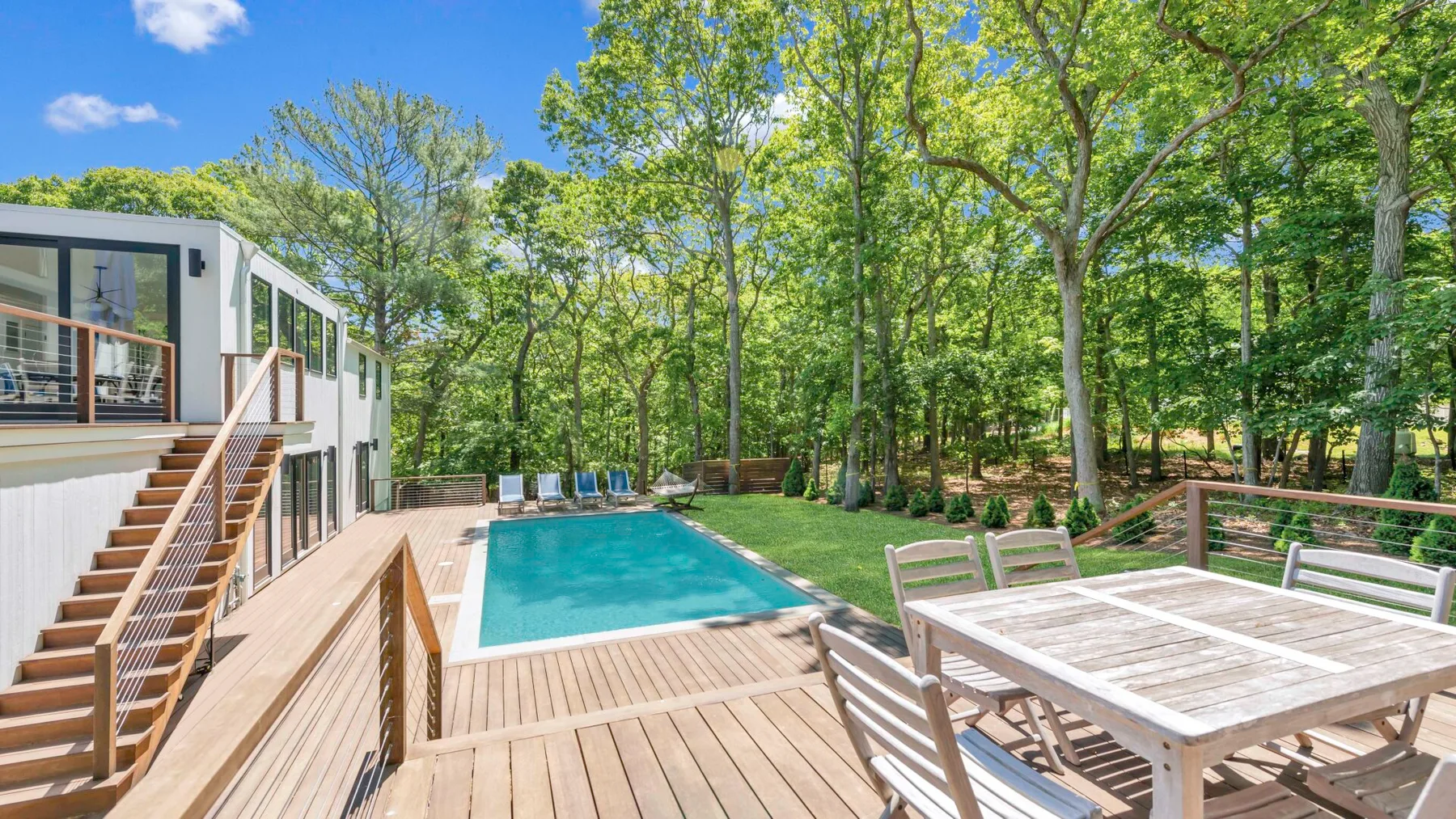 Un patio arbolado cuenta con una piscina, una terraza de madera y muebles para comer al aire libre.