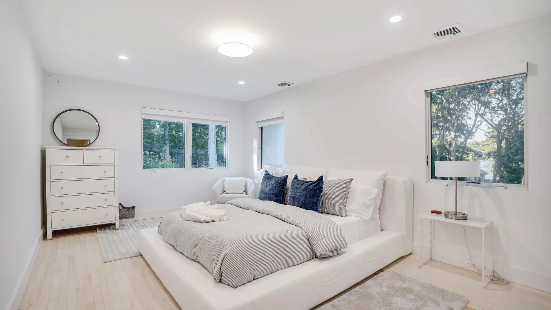 Un dormitorio luminoso con una cama grande, ropa de cama neutra y ventanas con vista a los árboles.
