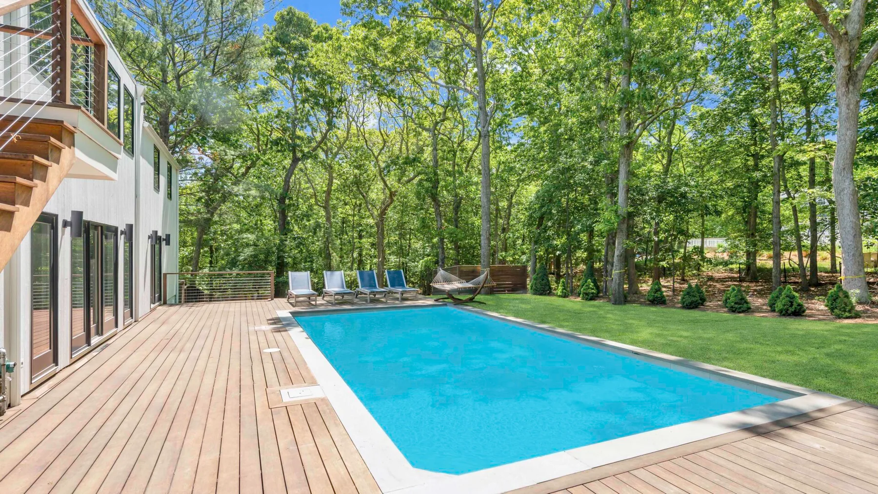 Una piscina junto a una terraza de madera, con árboles y césped cerca.