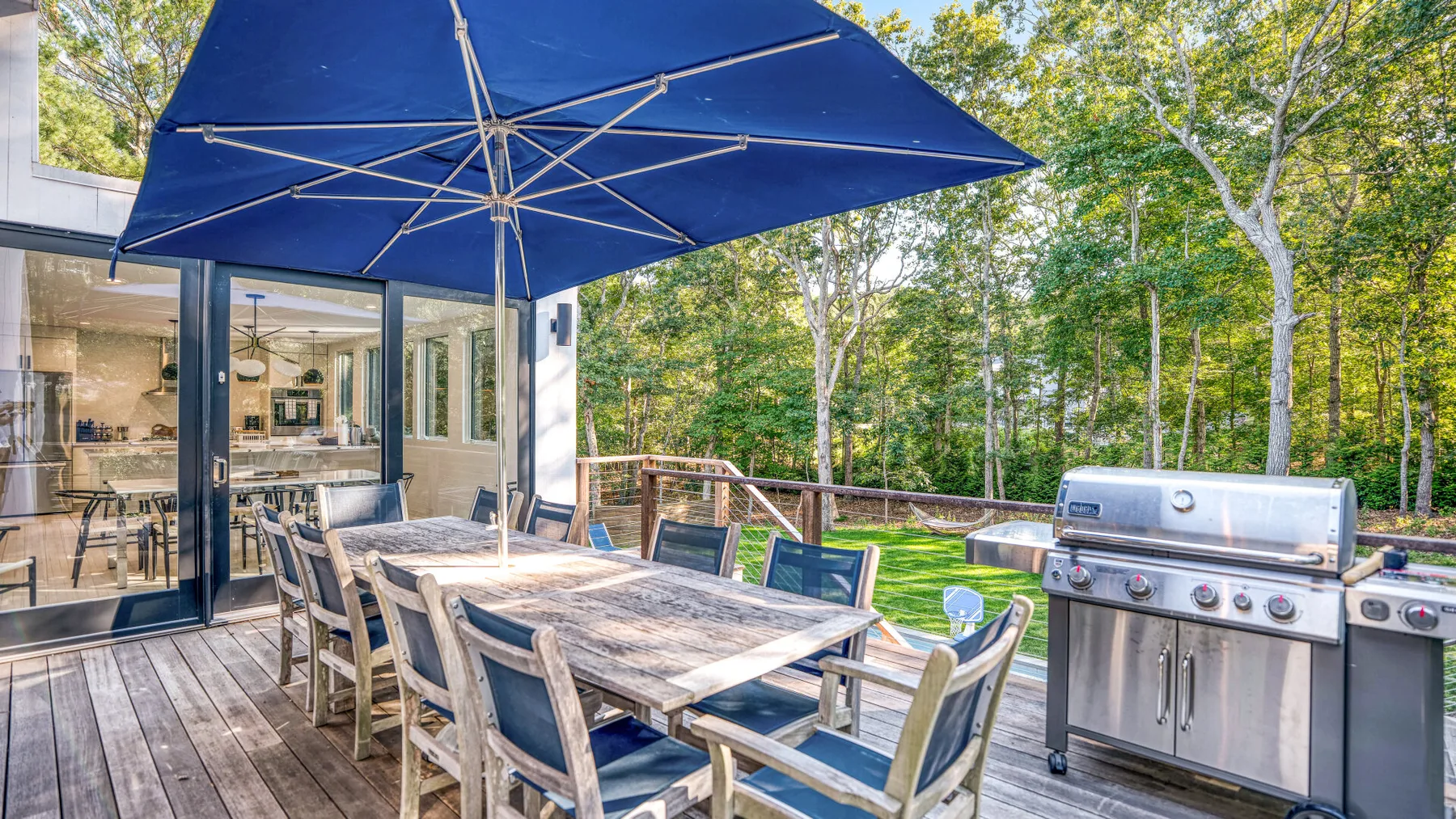 Un comedor exterior de madera está bajo una gran sombrilla azul junto a la casa.