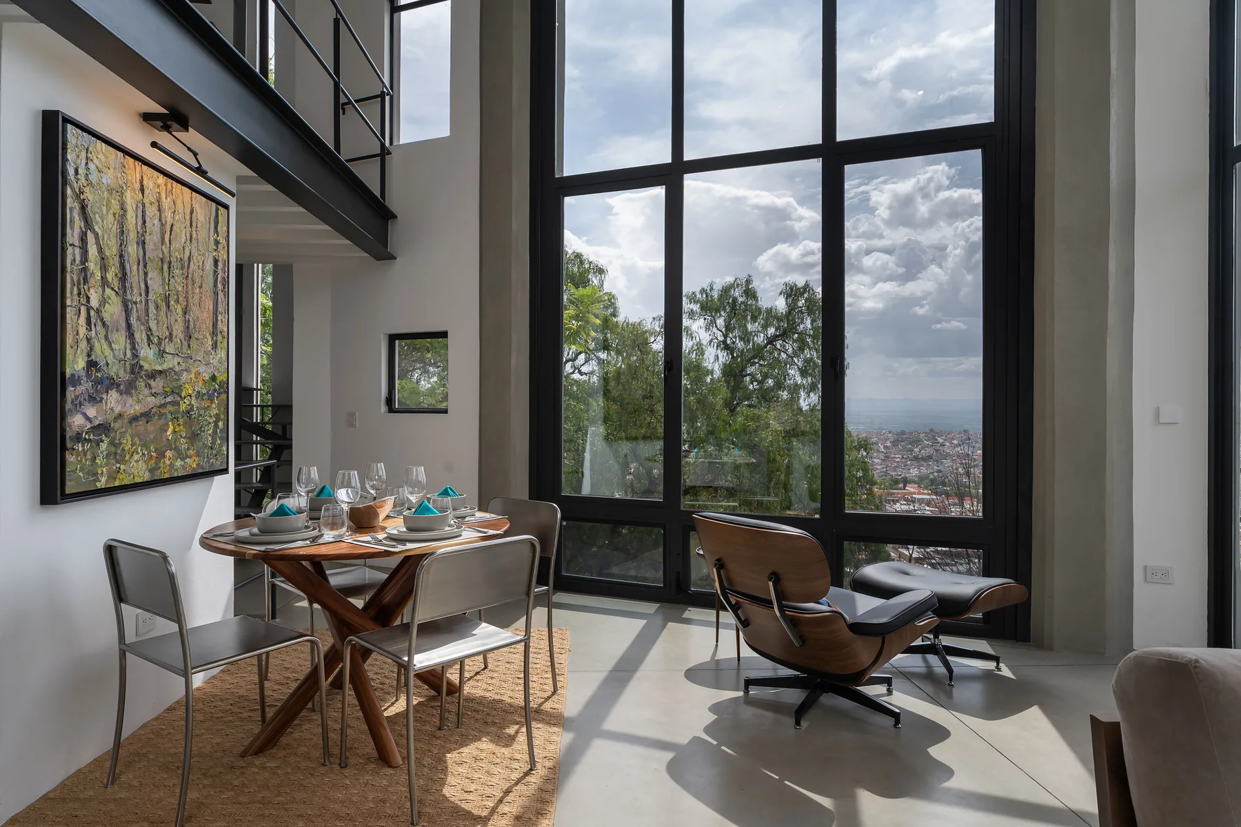A bright dining and sitting area features a table beside tall windows overlooking trees and the city.