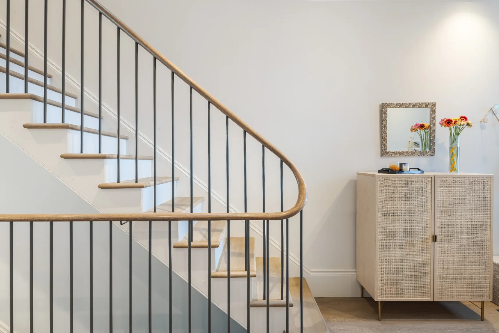 Una escalera con barandilla oscura está junto a un mueble con flores encima.