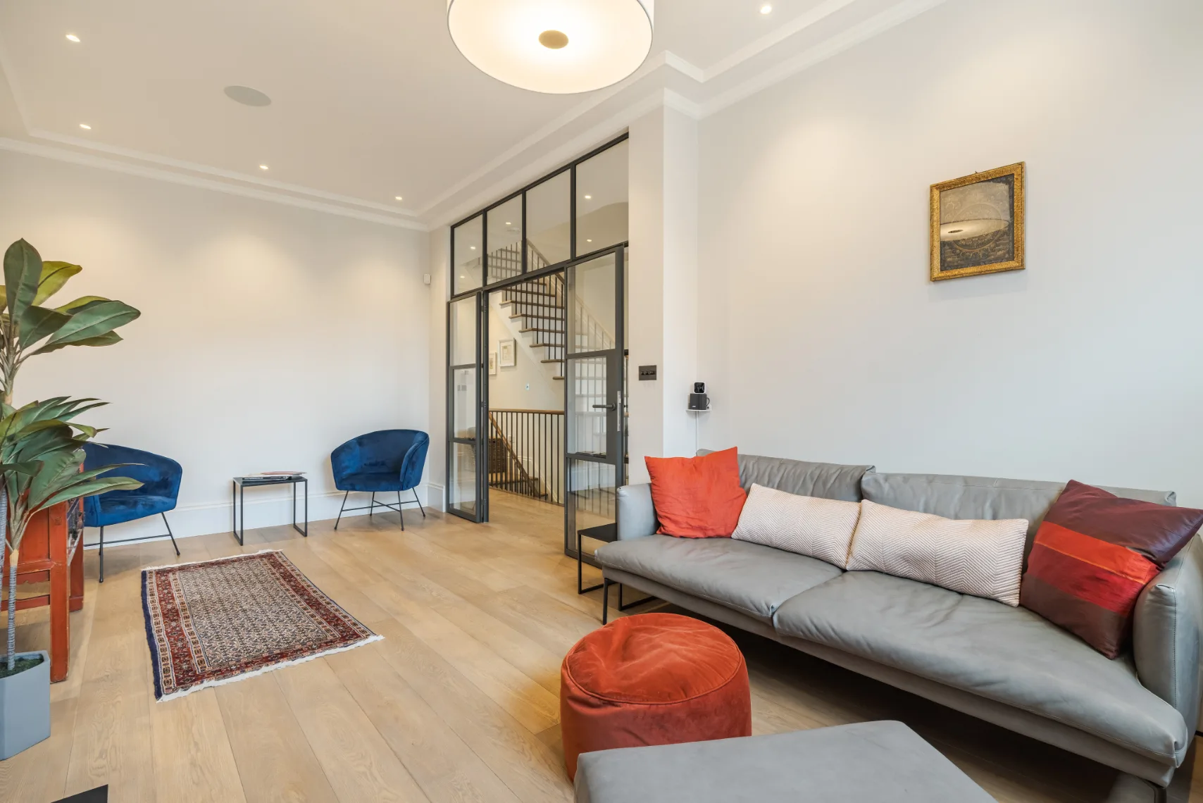 Una sala luminosa con un sofá gris largo y un rincón de asientos junto a la mampara de vidrio.