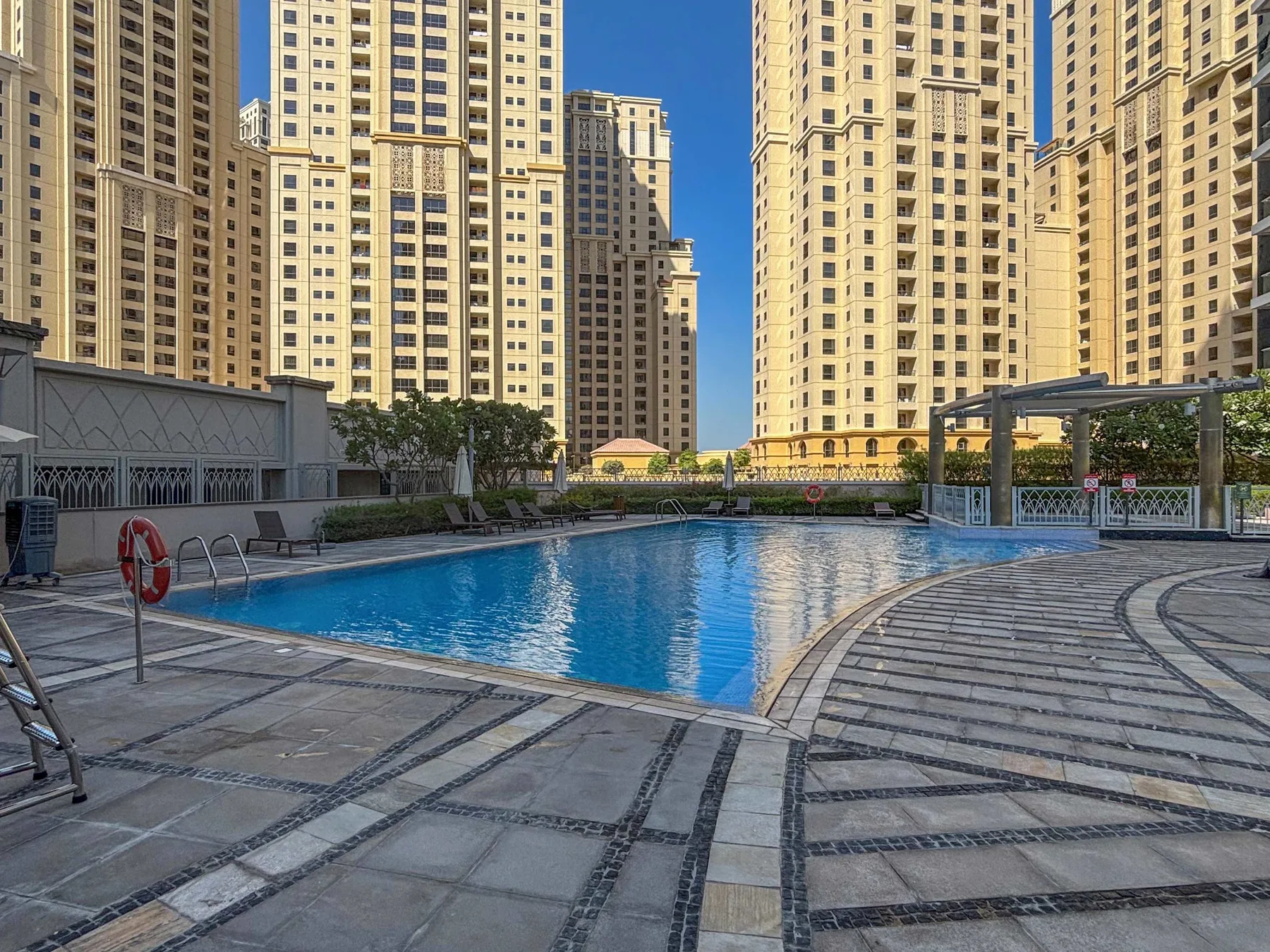Una piscina al aire libre se encuentra entre edificios residenciales altos, con zonas pavimentadas alrededor.