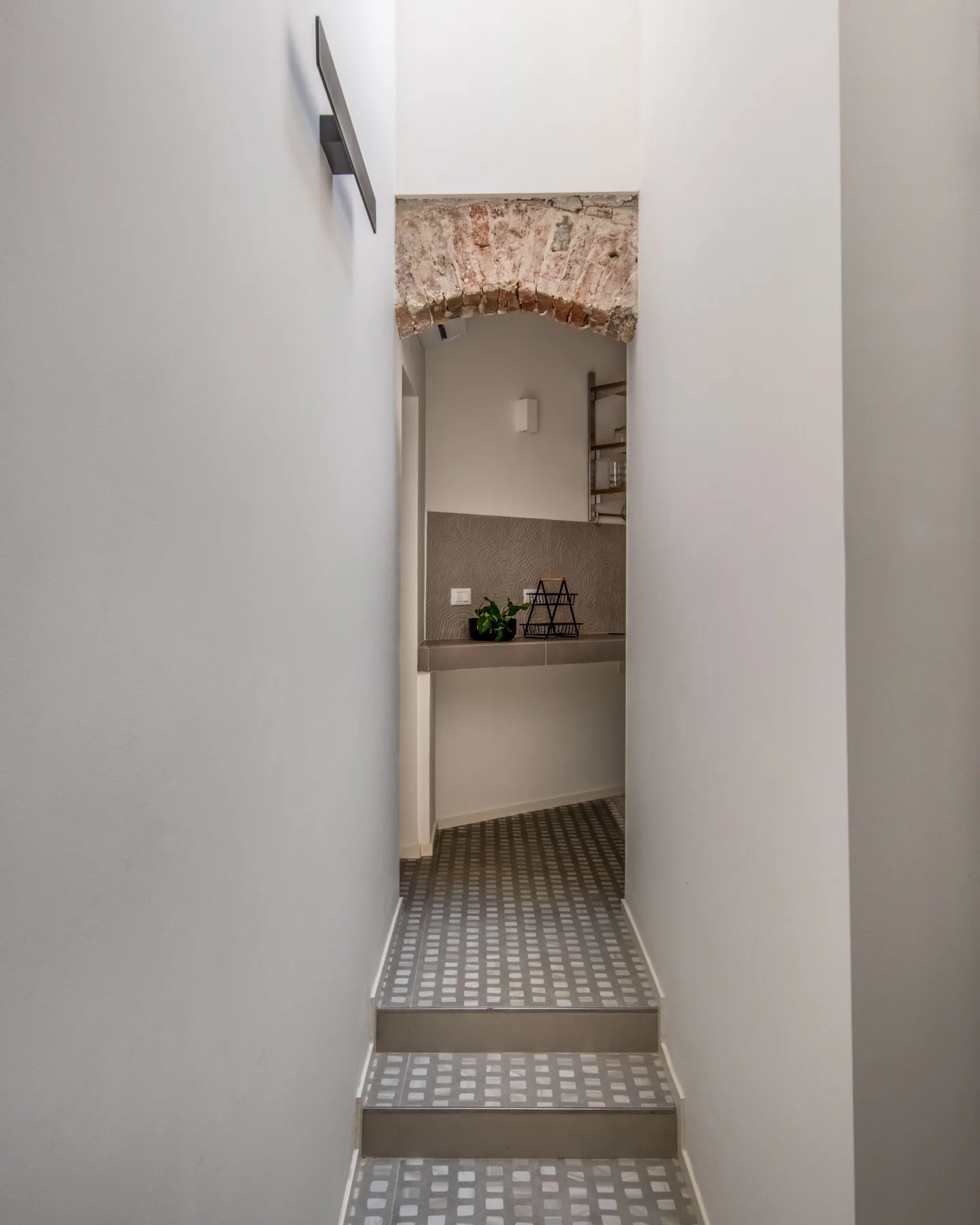 A narrow hallway with patterned tile flooring leads to a small counter beneath a brick arch.
