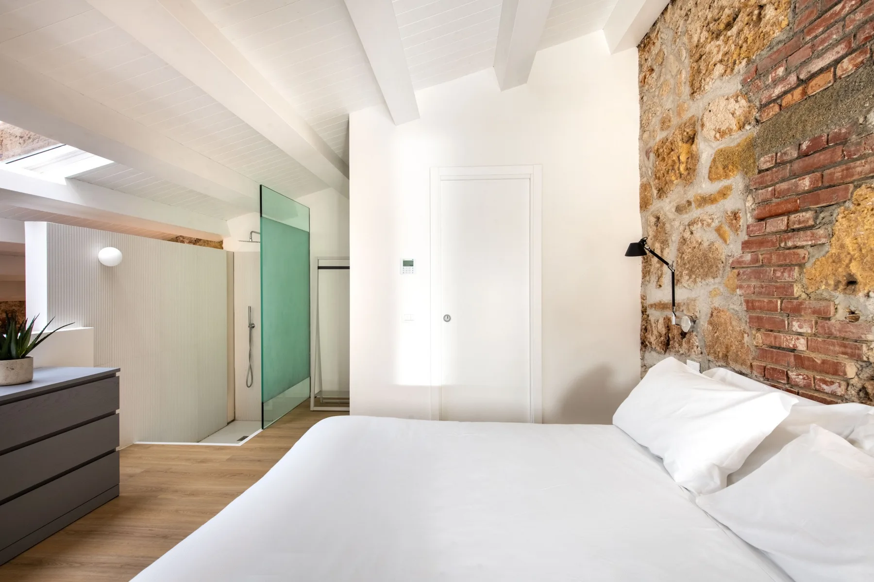 A bright bedroom with a white bed beside an exposed brick and stone wall.