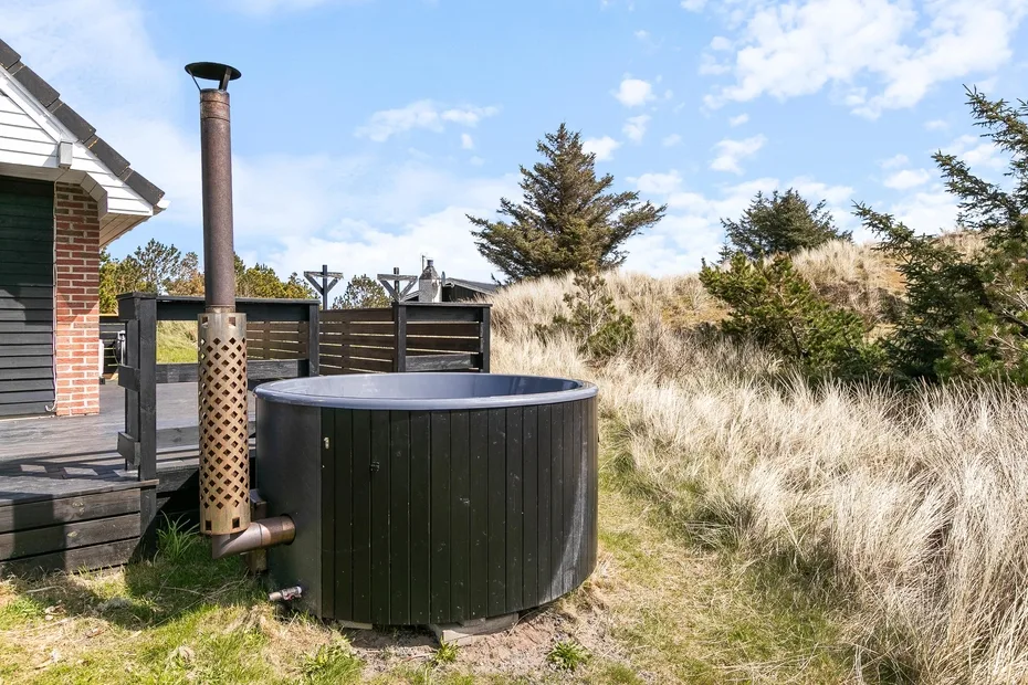 Una bañera de hidromasaje exterior está junto a una terraza de madera entre dunas cubiertas de hierba.