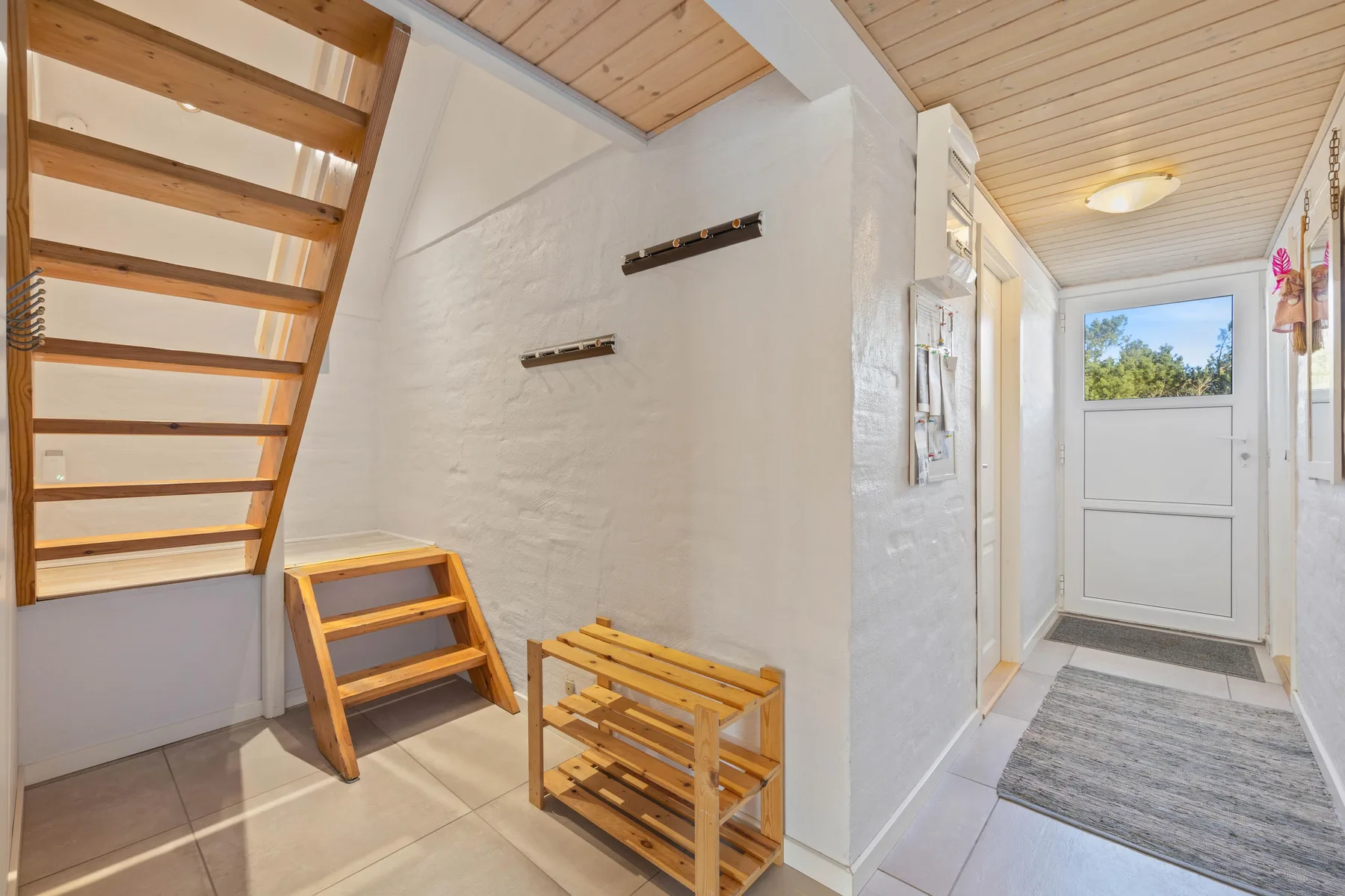Una entrada luminosa tiene una puerta blanca y una escalera de madera.