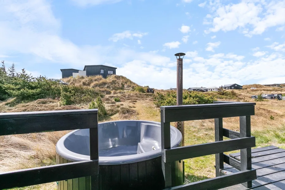Una bañera de hidromasaje exterior está junto a una terraza de madera, frente a dunas cubiertas de hierba.
