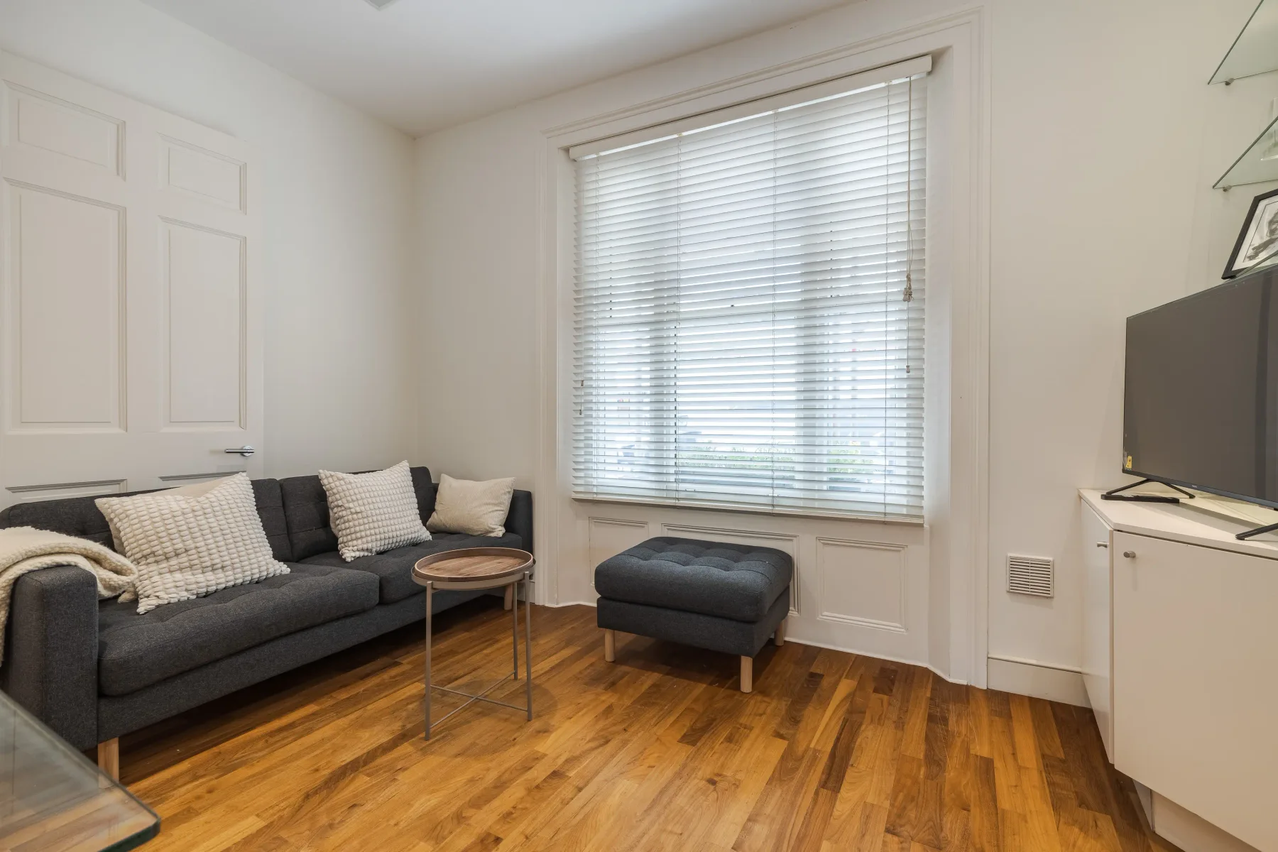 A living room with dark seating, a window with blinds, and a wooden floor.