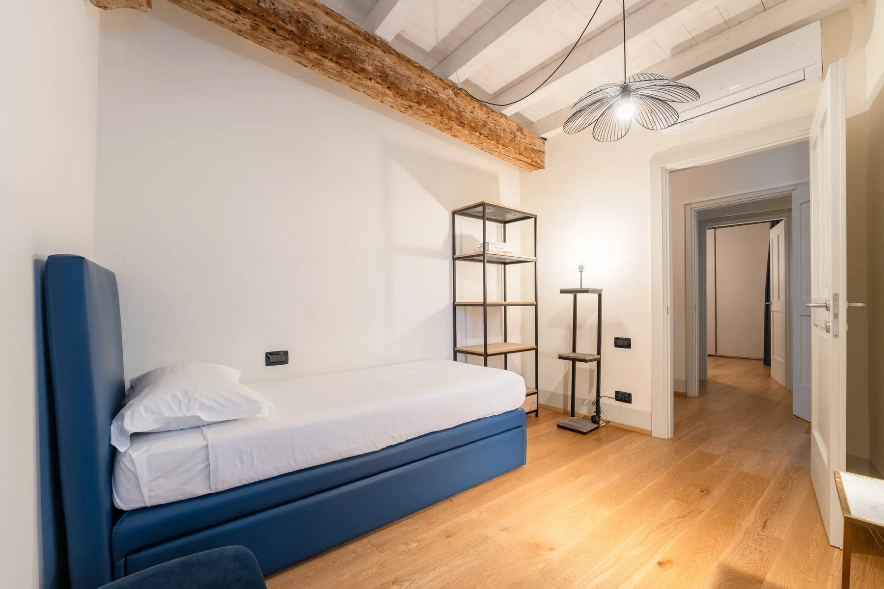 Un dormitorio con una cama individual, estanterías abiertas y una puerta hacia el pasillo.