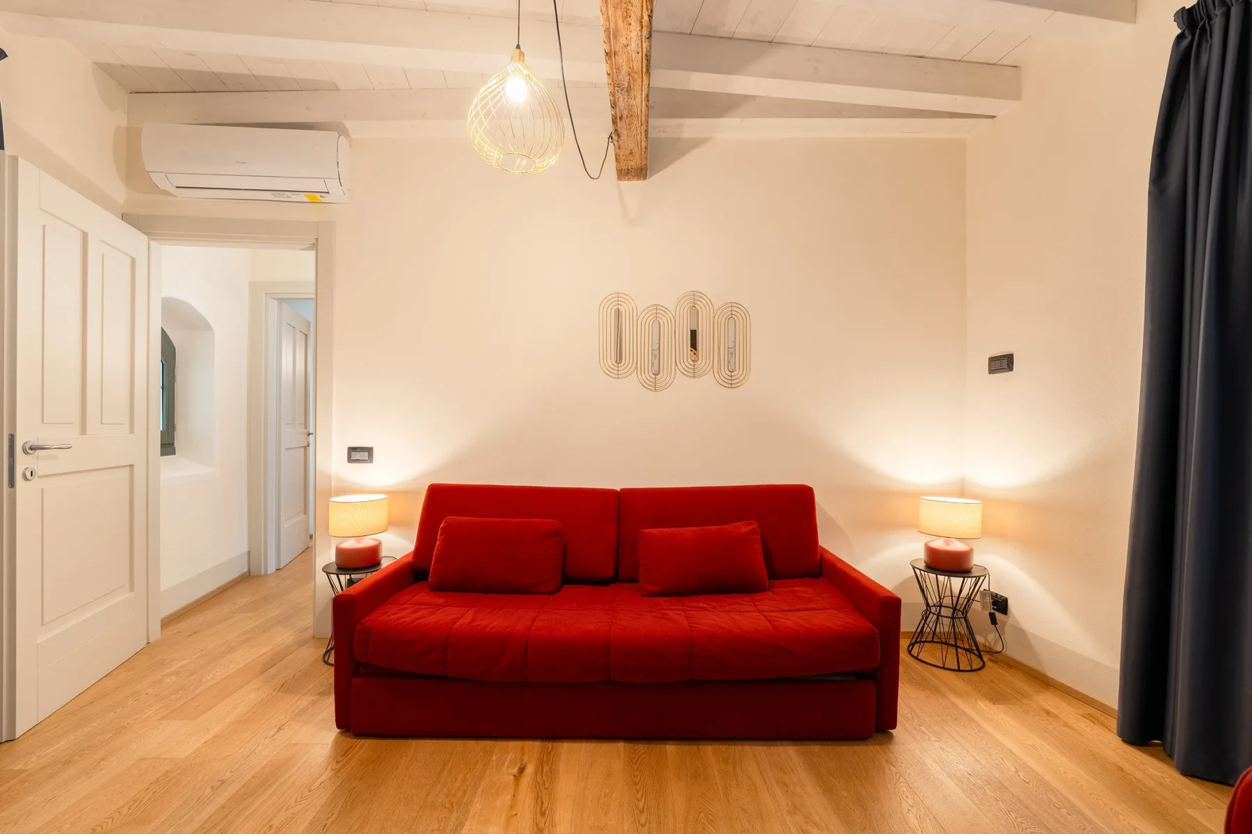 Una sala luminosa con un sofá rojo, lámparas cálidas y suelo de madera.