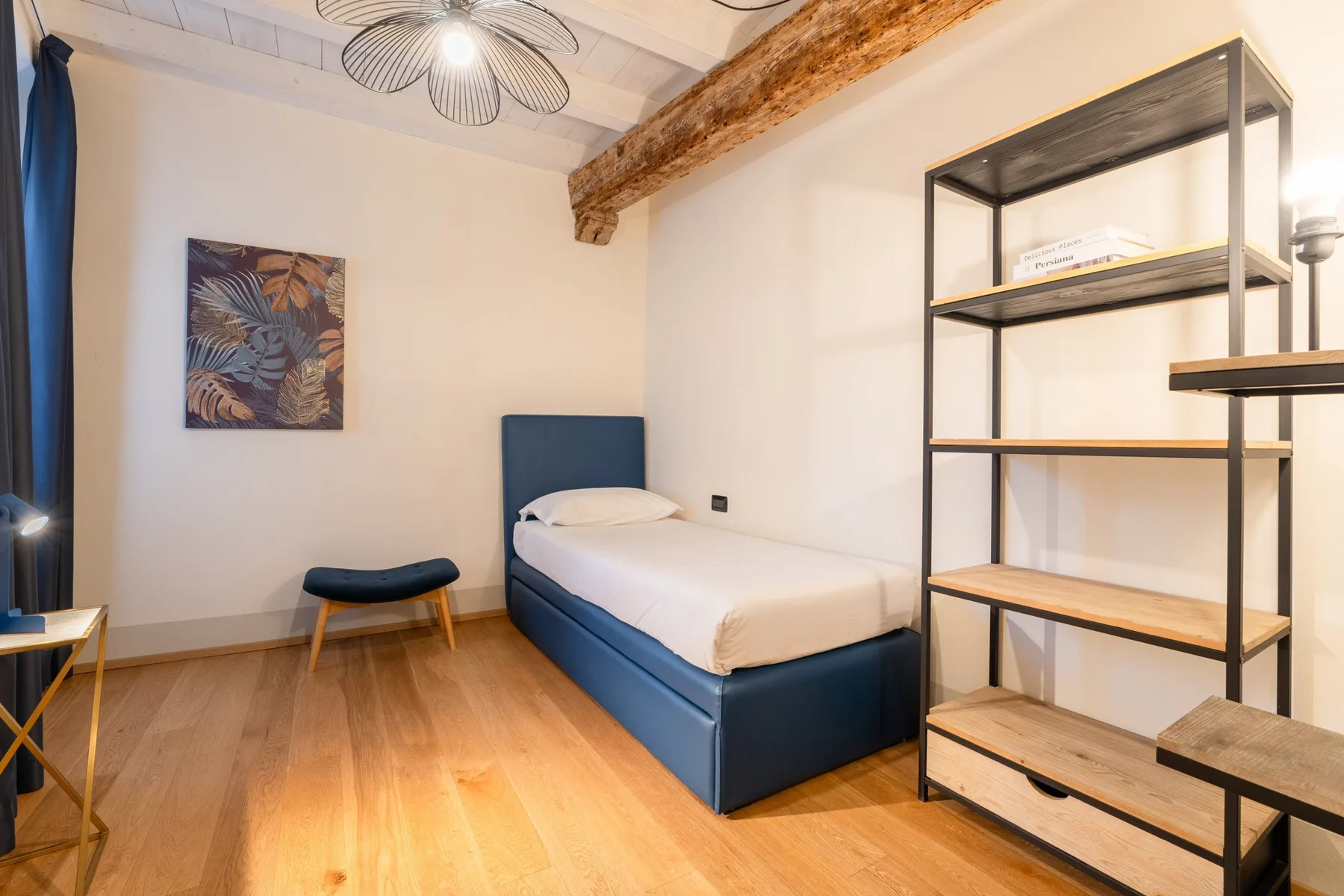 Un dormitorio con una cama azul, un pequeño taburete y estanterías abiertas.