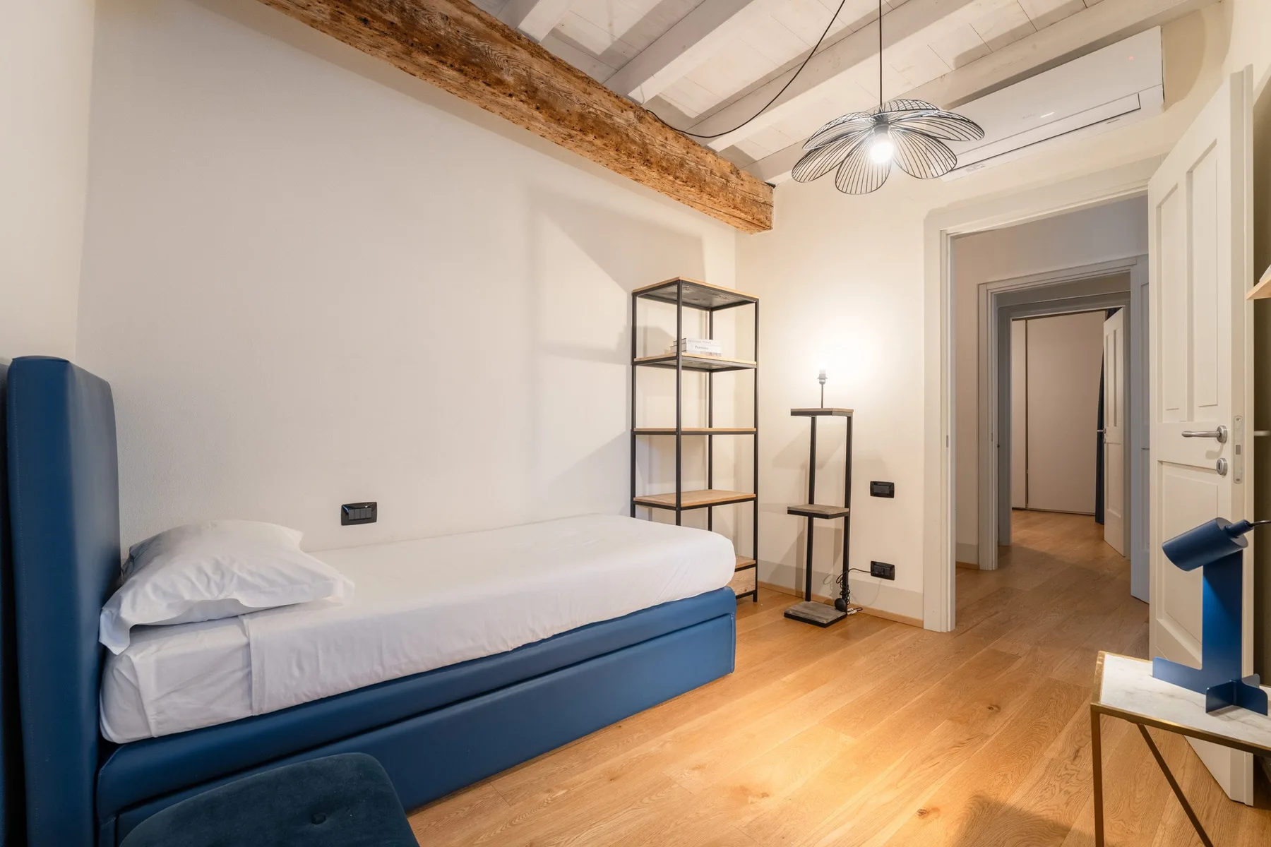 Un dormitorio con una cama azul, estanterías abiertas y una puerta al pasillo.