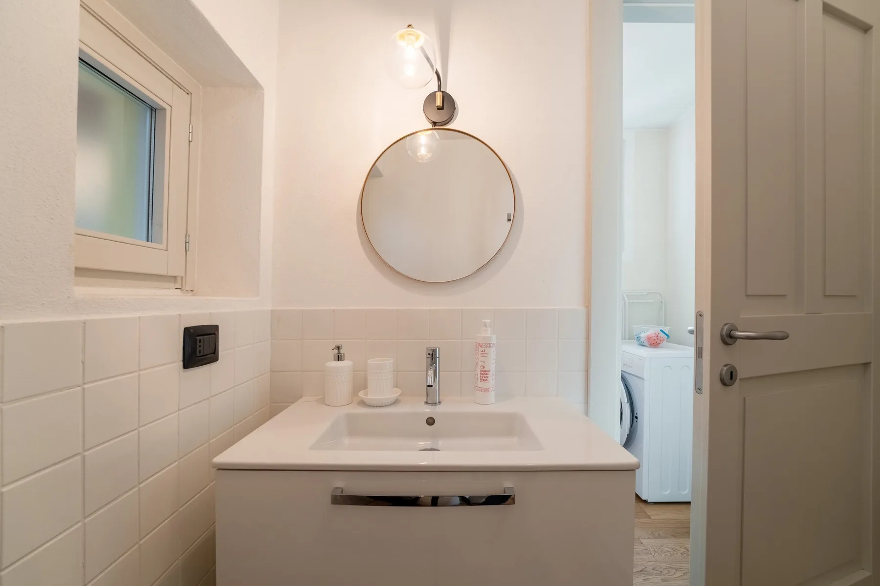 Un tocador de baño luminoso con un espejo redondo y una ventana pequeña.
